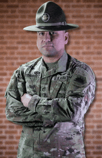 A Man In A U.s. Army Uniform Stands With His Arms Crossed In Front Of A Brick Wall GIF