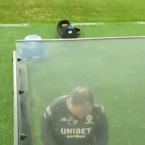 A Man In A Unibet Jersey Is Standing On A Soccer Field Behind A Glass Barrier . GIF