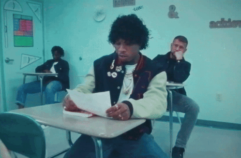 A Man In A Varsity Jacket Is Sitting At A Desk Reading A Piece Of Paper GIF