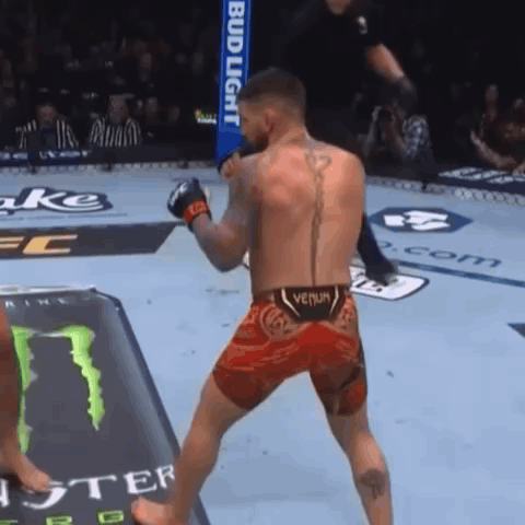 A Man In A Venum Shorts Stands In Front Of A Bud Light Sign GIF
