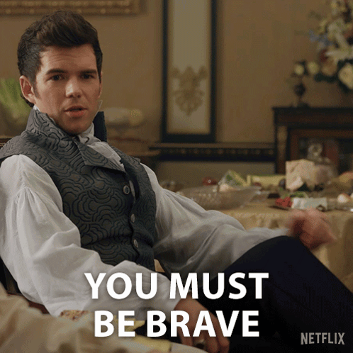 A Man In A Vest Sits In Front Of A Table With The Words " You Must Be Brave " On It GIF