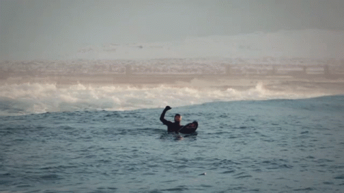 A Man In A Wetsuit Is Floating In The Ocean With His Hand Up GIF