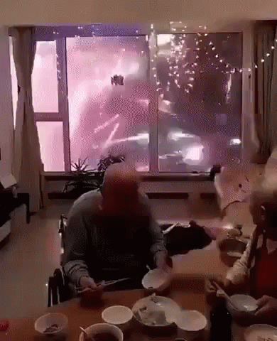 A Man In A Wheelchair Sits At A Table With Bowls And Plates In Front Of A Window Watching Fireworks GIF