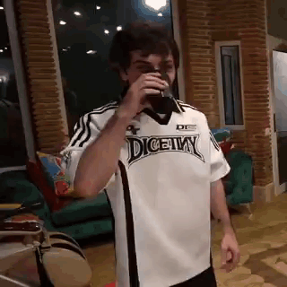 A Man In A White And Black Soccer Jersey Is Drinking From A Cup In A Living Room . GIF