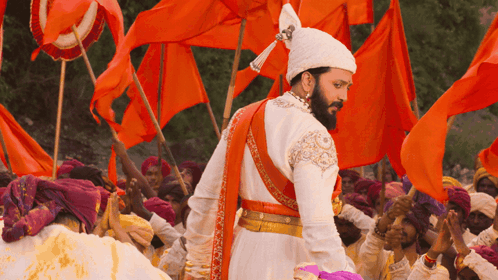 A Man In A White And Orange Costume Stands In Front Of A Crowd Of People Holding Orange Flags GIF