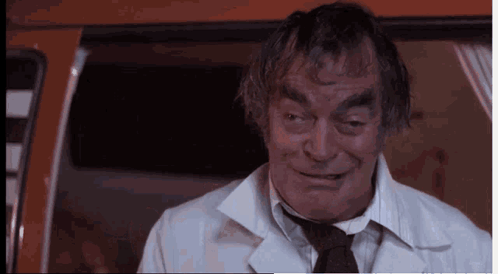 A Man In A White Coat And Tie Is Smiling And Looking At The Camera GIF