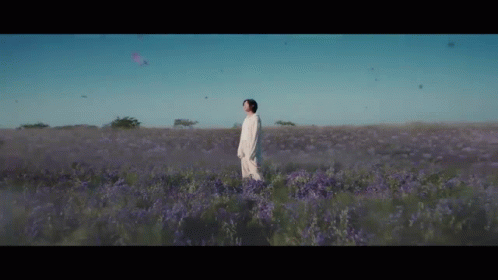 A Man In A White Dress Is Standing In A Field Of Purple Flowers . GIF