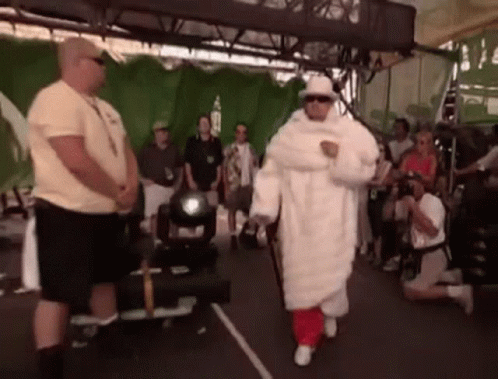 A Man In A White Fur Coat And Hat Is Walking In Front Of A Crowd . GIF