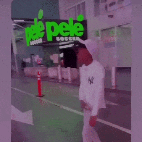 A Man In A White Hat Is Standing In Front Of A Building With A Green Sign That Says Soccer . GIF