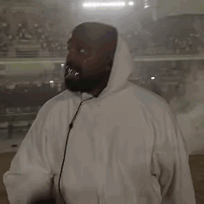 A Man In A White Hoodie Is Holding A Microphone In Front Of A Crowd . GIF