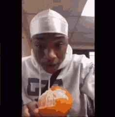 A Man In A White Hoodie Is Holding An Orange In His Hand . GIF