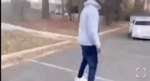A Man In A White Hoodie Is Walking Down A Street Next To A Car . GIF