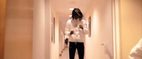 A Man In A White Jacket Is Walking Down A Hallway With Stairs . GIF