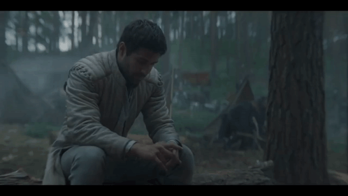 A Man In A White Jacket Sits In The Woods With His Hands On His Knees GIF