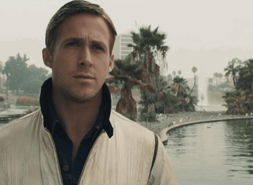 A Man In A White Jacket Stands In Front Of A Pond With Palm Trees In The Background GIF