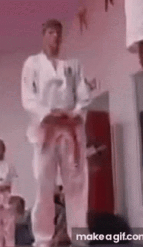 A Man In A White Karate Uniform Is Standing In A Room With His Belt Around His Waist . GIF