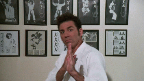 A Man In A White Karate Uniform Stands In Front Of A Wall Of Framed Pictures Of Karate Fighters GIF