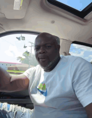 A Man In A White Nike Shirt Is Sitting In A Car GIF