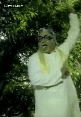 A Man In A White Robe And Glasses Is Standing In Front Of A Tree With His Arms In The Air . GIF