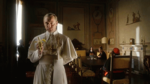 A Man In A White Robe Holds A Banana In A Room GIF