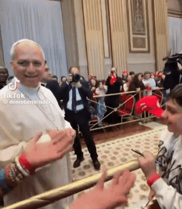 A Man In A White Robe Is Shaking Hands With A Boy In A Red Hat In Front Of A Crowd . GIF