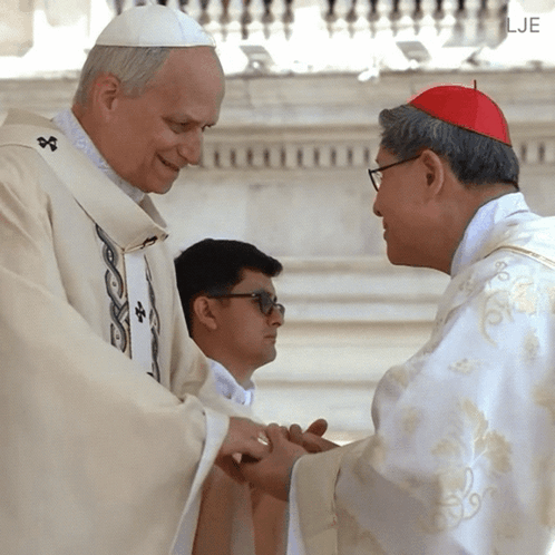 A Man In A White Robe Is Shaking Hands With A Man Wearing A Red Hat GIF