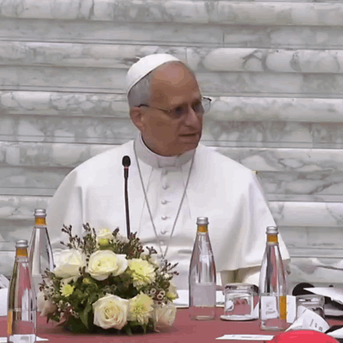 A Man In A White Robe Is Sitting At A Table With Bottles Of Water And Flowers GIF