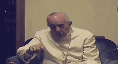 A Man In A White Robe Is Sitting In A Chair With A Cross On His Neck . GIF
