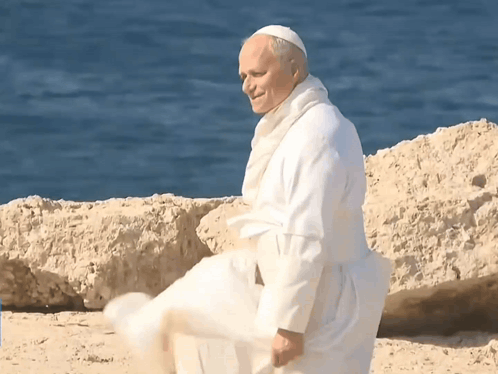 A Man In A White Robe Is Standing On A Beach Near The Water GIF