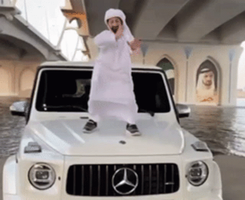 A Man In A White Robe Is Standing On The Hood Of A White Mercedes GIF