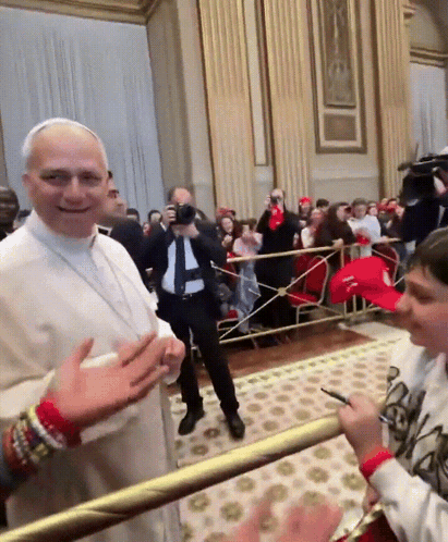 A Man In A White Robe Stands In Front Of A Crowd While A Boy In A Red Hat Stands Behind Him GIF