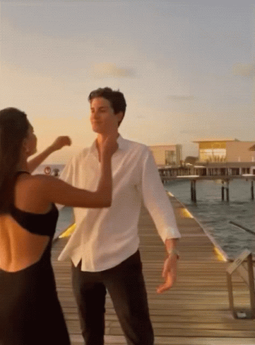 A Man In A White Shirt And A Woman In A Black Dress Are Dancing On A Dock GIF