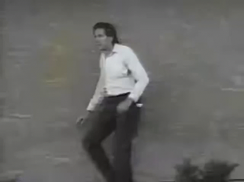 A Man In A White Shirt And Black Pants Is Standing In Front Of A Brick Wall . GIF