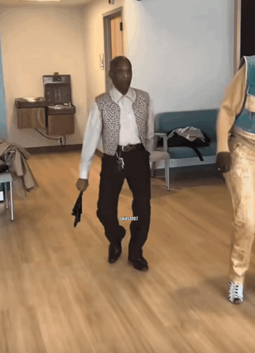 A Man In A White Shirt And Black Pants Is Walking In A Hallway With A Watermark That Says Shills1987 GIF