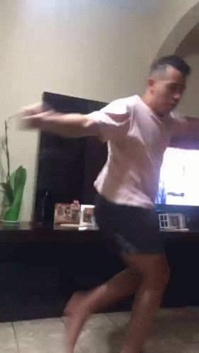 A Man In A White Shirt And Black Shorts Is Dancing In Front Of A Tv GIF