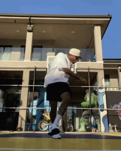 A Man In A White Shirt And Black Shorts Stands On A Basketball Court In Front Of A House GIF