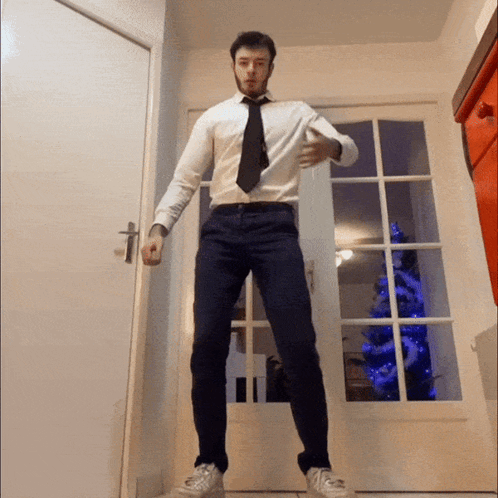 A Man In A White Shirt And Black Tie Is Standing In Front Of A Christmas Tree GIF
