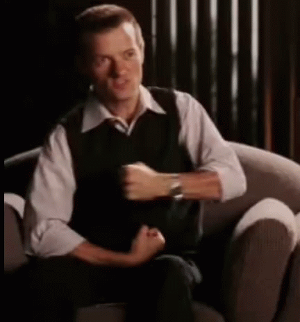 A Man In A White Shirt And Black Vest Is Sitting In A Chair . GIF