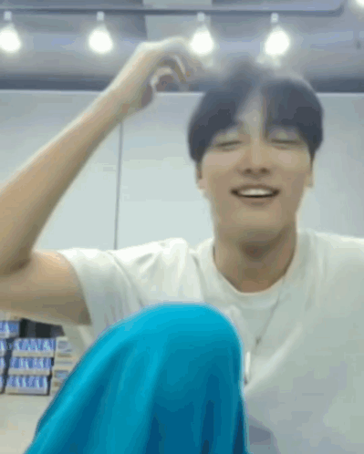 A Man In A White Shirt And Blue Pants Is Sitting On The Floor With His Legs Crossed And Smiling . GIF