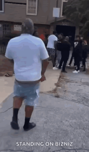 A Man In A White Shirt And Blue Shorts Is Standing On A Sidewalk In Front Of A Group Of People . GIF