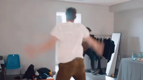 A Man In A White Shirt And Brown Pants Is Dancing In A Room In Front Of A Mirror . GIF