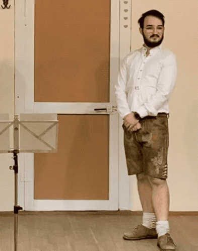 A Man In A White Shirt And Brown Shorts Stands In Front Of A Music Stand GIF