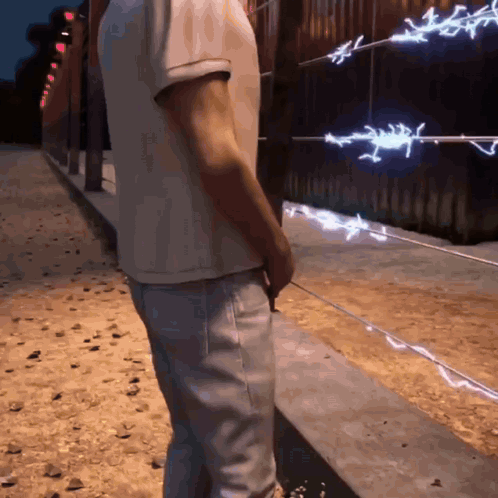 A Man In A White Shirt And Jeans Stands In Front Of A Barbed Wire Fence GIF