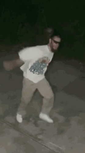 A Man In A White Shirt And Khaki Pants Is Dancing On A Sidewalk At Night . GIF