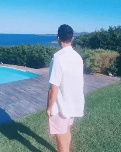 A Man In A White Shirt And Pink Shorts Is Standing In Front Of A Swimming Pool GIF