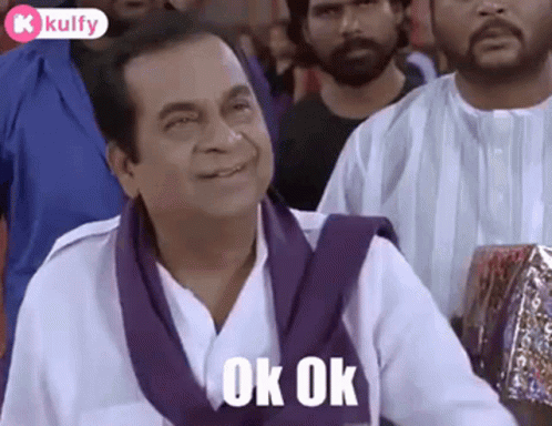 A Man In A White Shirt And Purple Scarf Is Sitting In A Crowd Of People And Smiling . GIF