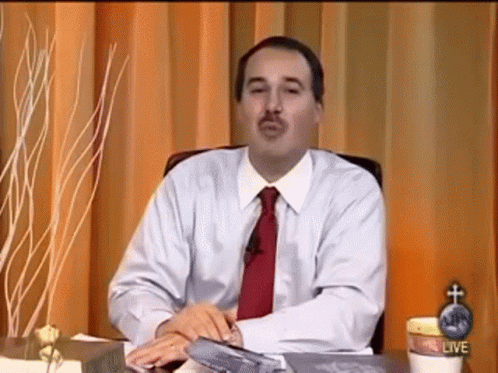 A Man In A White Shirt And Red Tie Is Sitting At A Desk In Front Of A Curtain . GIF