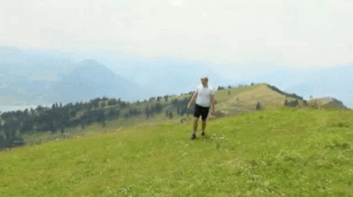 A Man In A White Shirt And Shorts Is Standing On Top Of A Grassy Hill . GIF