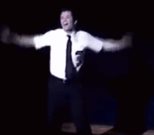 A Man In A White Shirt And Tie Is Dancing On A Stage In The Dark . GIF