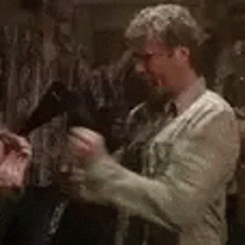 A Man In A White Shirt And Tie Is Standing In A Dark Room Holding A Gun . GIF